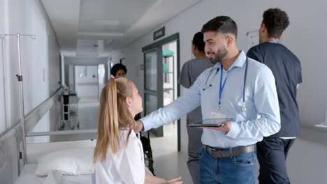 Biracial-male-doctor-and-caucasian-girl-patient-sitting-on-bed-talking-in-hospital,-slow-motion