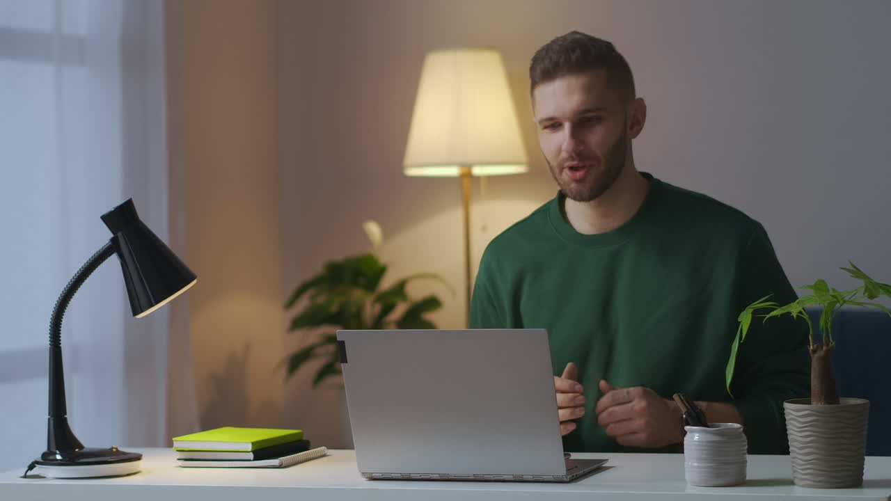handsome man is communicating by video chat by laptop smiling and gesticulating looking at screen sitting at home at evening