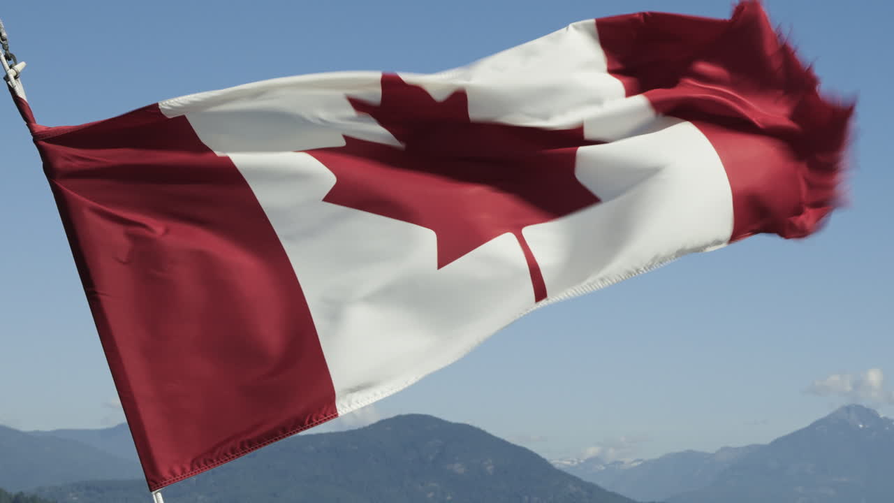 la bandera canadiense ondeando en el viento contra un cielo azul con montañas verdes en la distancia