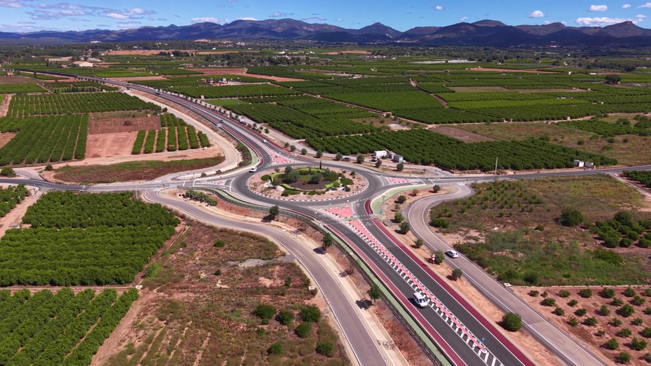 Gran rotonda de autopista rodeada de campos agrícolas verdes, huertos y viñedos, con una cordillera visible bajo un cielo azul en la España rural.