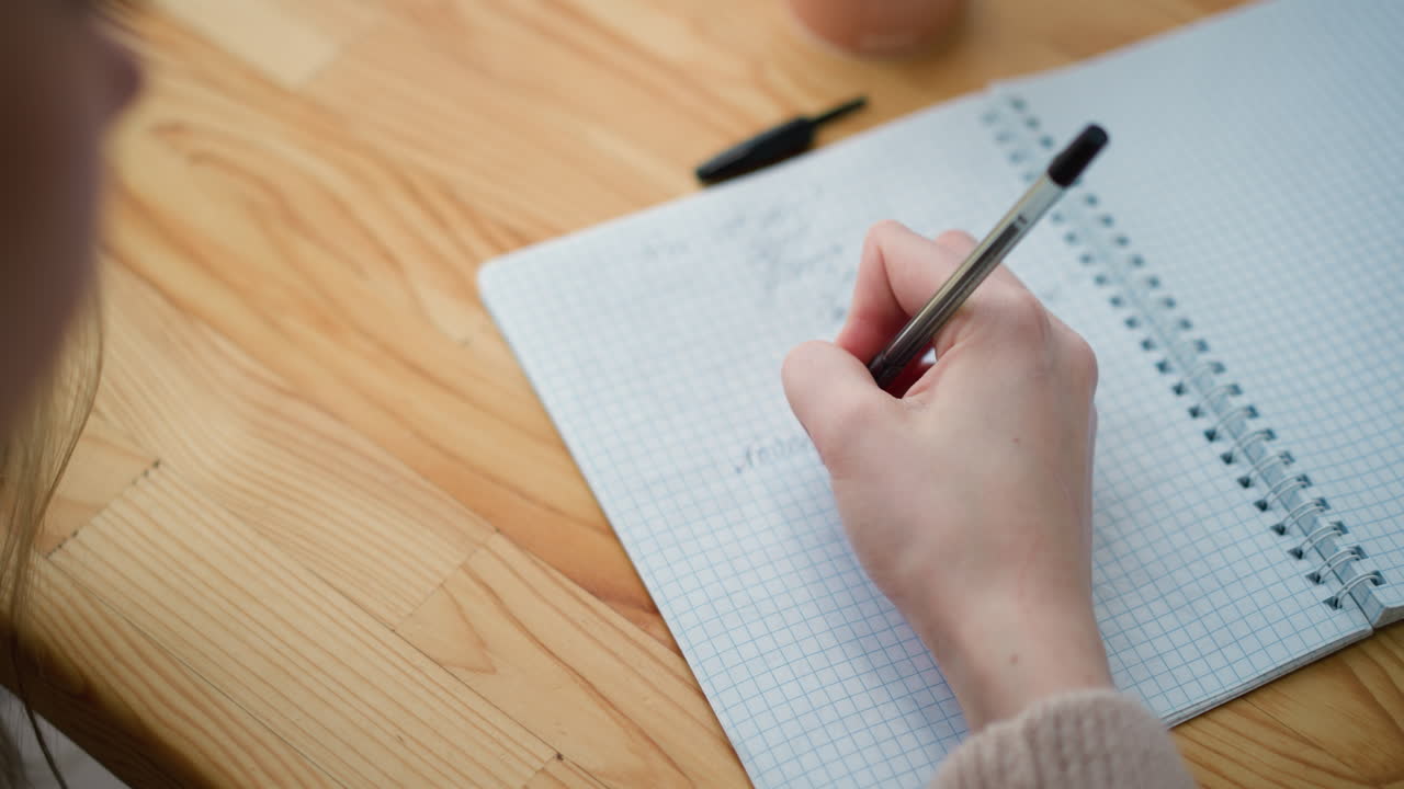 Close-up of hand holding pen, writing in notebook with partial view of coffee cup on table, focus on concentration, work, and productivity, soft background with grid pattern on paper