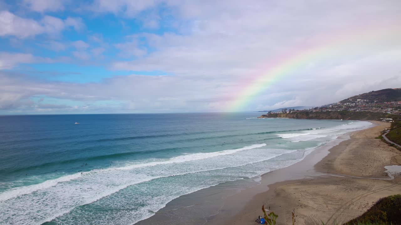 un hermoso lapso de tiempo de 4k desde el acantilado de una playa de dana point california justo después de que lloviera con un arco iris en el fondo mientras los surfistas navegan y las familias disfrutan de un día soleado de vacaciones