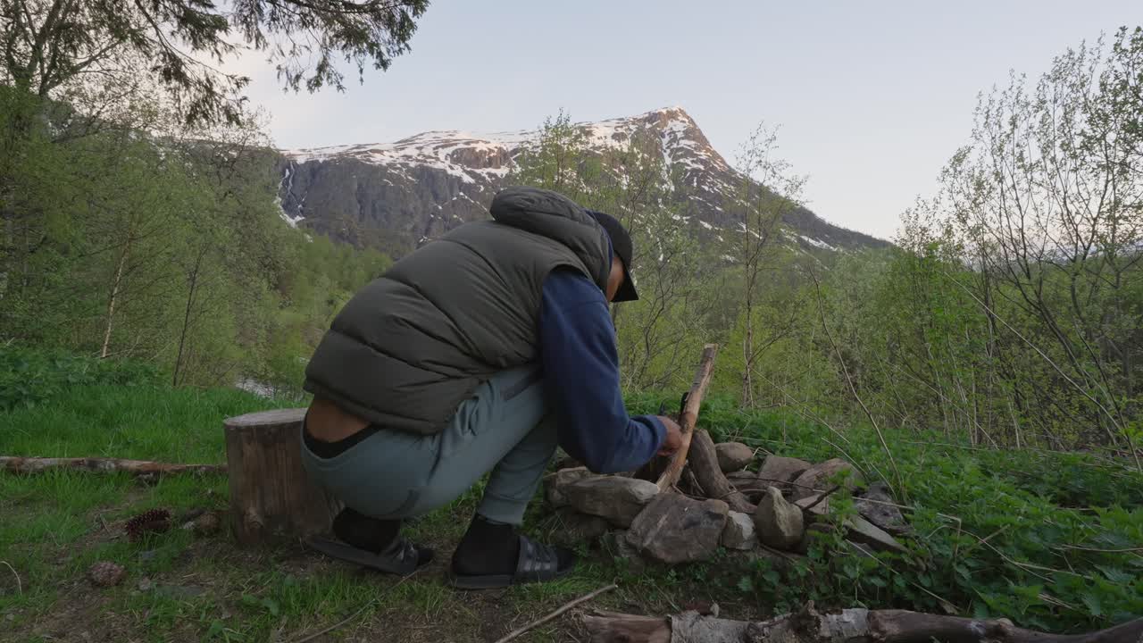 Uomo che accende un falò nella natura in un campeggio selvaggio con vista sulle montagne.
