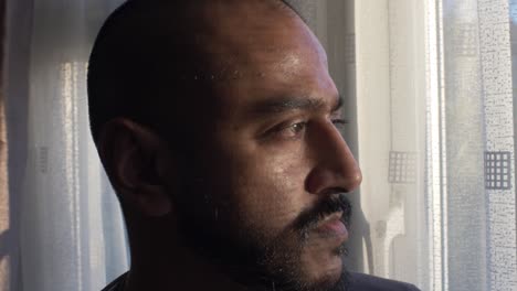 adult asian male in deep thought looking out to window with white curtains