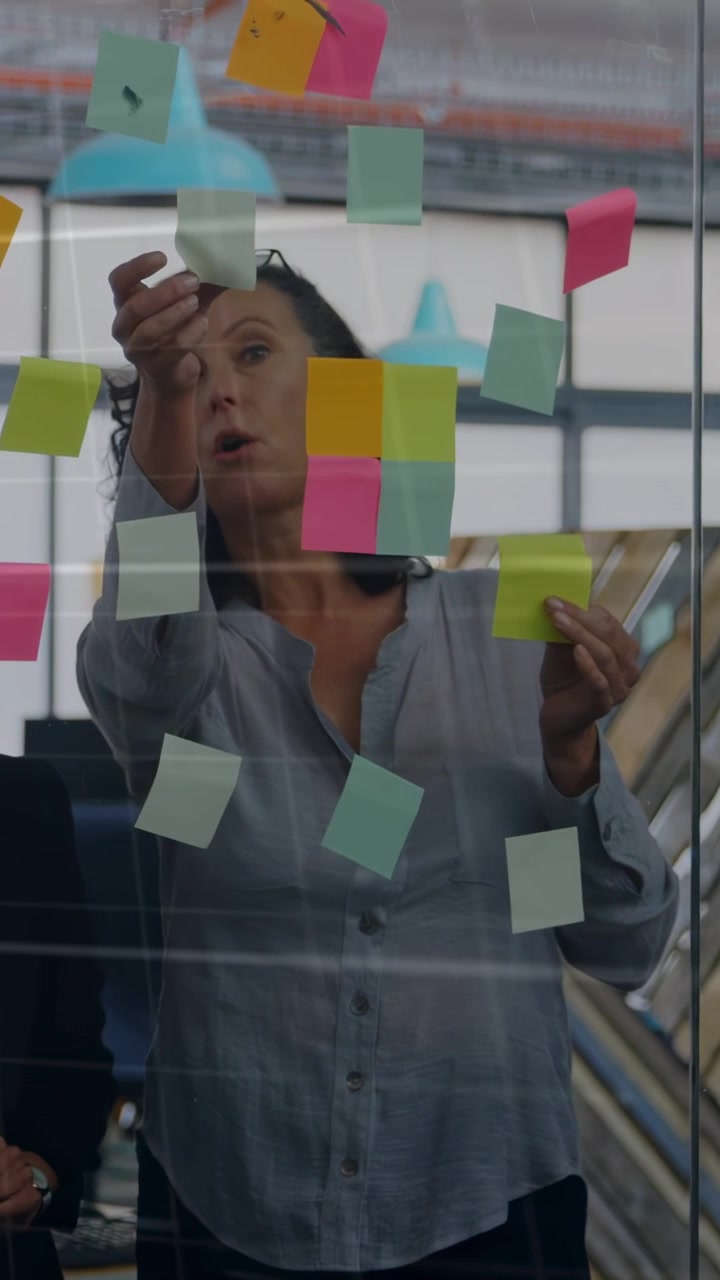 Vídeo vertical: Líder de escritório descolando nota verde, agrupando post-its no vidro, coordenando a equipe.