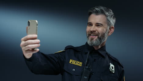 young joyful handsome policeman in uniform and with walkie talkie smiling and having videochat on the smartphone via web cam on the dark wall background