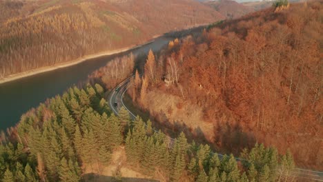 an aerial ascending drone view of two cars driving on a winding road alongside a lake in late autumn