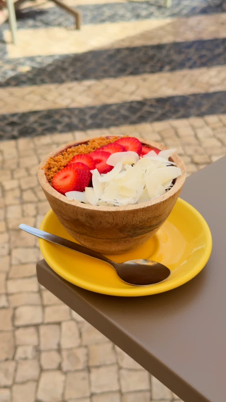 Tropical açai bowl topped with fresh strawberries, coconut flakes and paçoca, served in a wooden bowl on a yellow plate outdoors