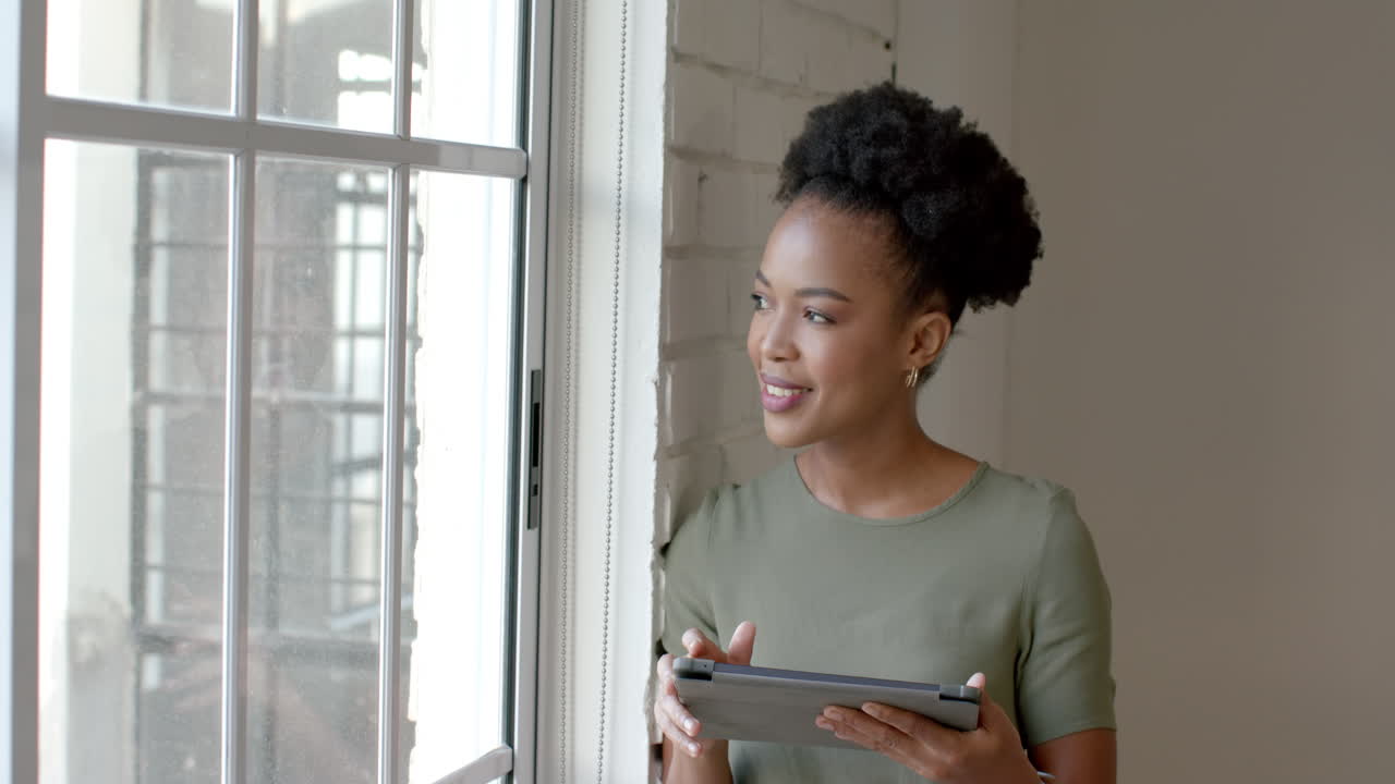 A young African American woman holds a tablet near a window, with copy space
