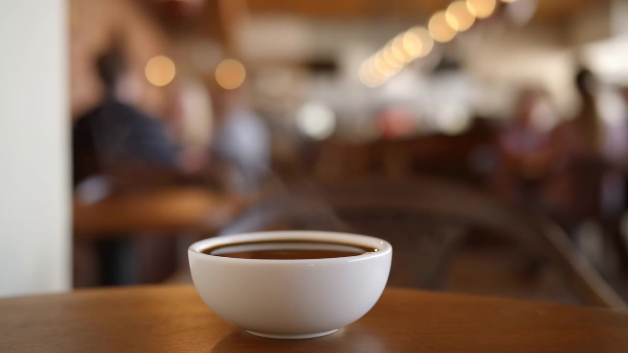 Tazza di caffè bianca su tavolo di legno in un vivace caffè urbano, luci bokeh calde e avventori sfocati, vapore che sale.