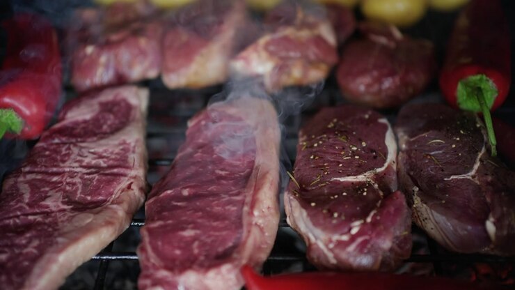 Daging sedang dipanggang di atas panggangan bersama terong dan paprika merah, dengan asap mengepul.