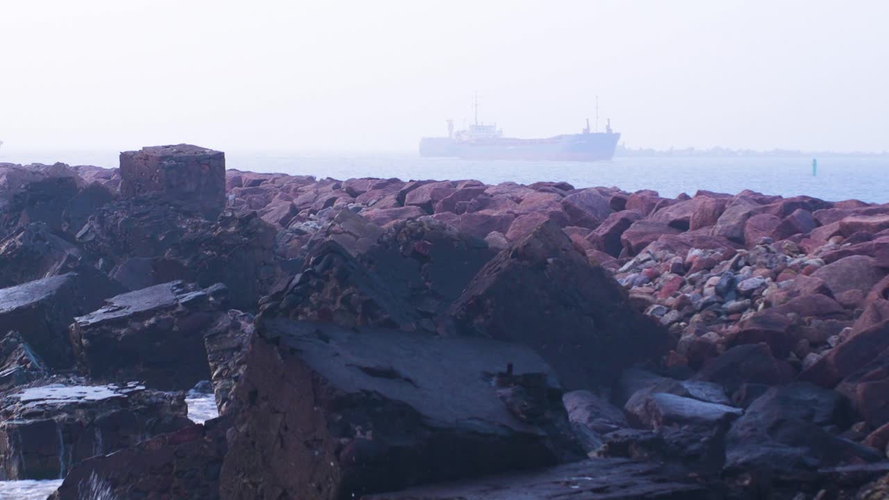 gran buque de carga entrando al puerto de liepaja en un día nublado, muelle de piedra en primer plano, olas salpicando, tiro medio distante