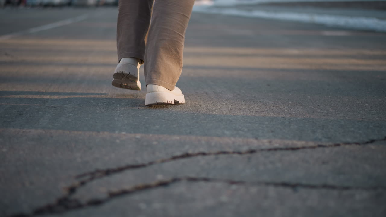 Back view of performer legs gently swaying on cracked pavement, wearing beige pants and white boots, casting long shadows under soft evening sunlight, evoking motion, and winter street rhythm