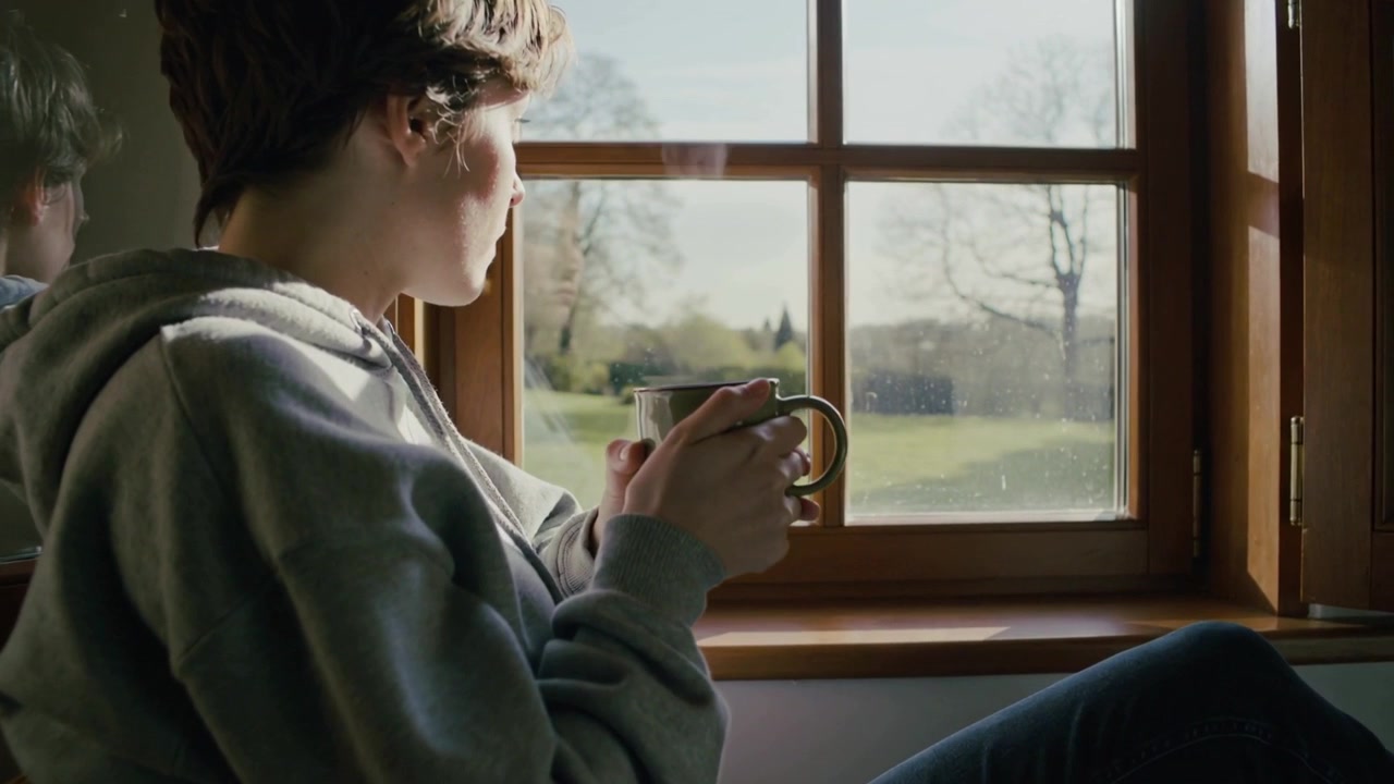 Adolescente con felpa grigia con cappuccio che sorseggia e tiene in mano una tazza fumante, guardando il cortile attraverso una finestra di legno, riflesso con la tazza