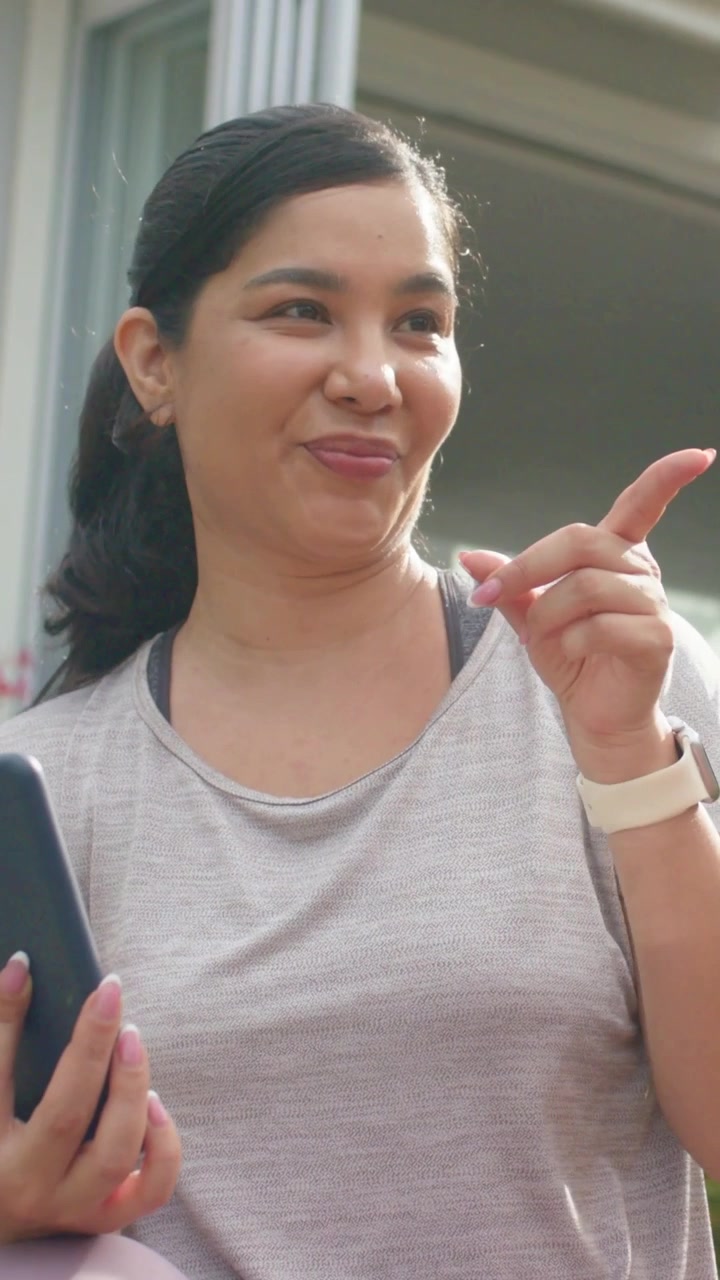 Video verticale: Una donna con maglietta rosa si sporge e indica, mentre una persona con maglietta grigia mostra il telefono e condivide una foto sul patio.