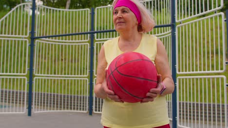 Mujer-Mayor-Abuela-Atleta-Posando-Jugando-Con-Pelota-Al-Aire-Libre-En-La-Cancha-De-Baloncesto