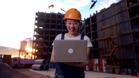 trabajadora asiática con gafas de protección y casco de seguridad sonriendo y señalando una computadora portátil en su mano mientras está de pie en el sitio de construcción