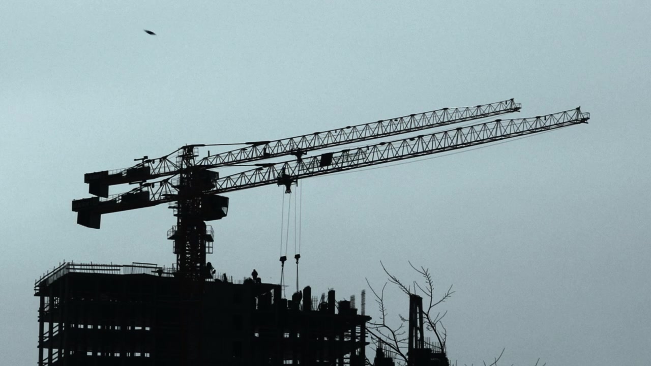 A large tower crane stands in silhouette over an unfinished building, with long lattice booms stretching across a pale hazy sky. Dark industrial forms line
