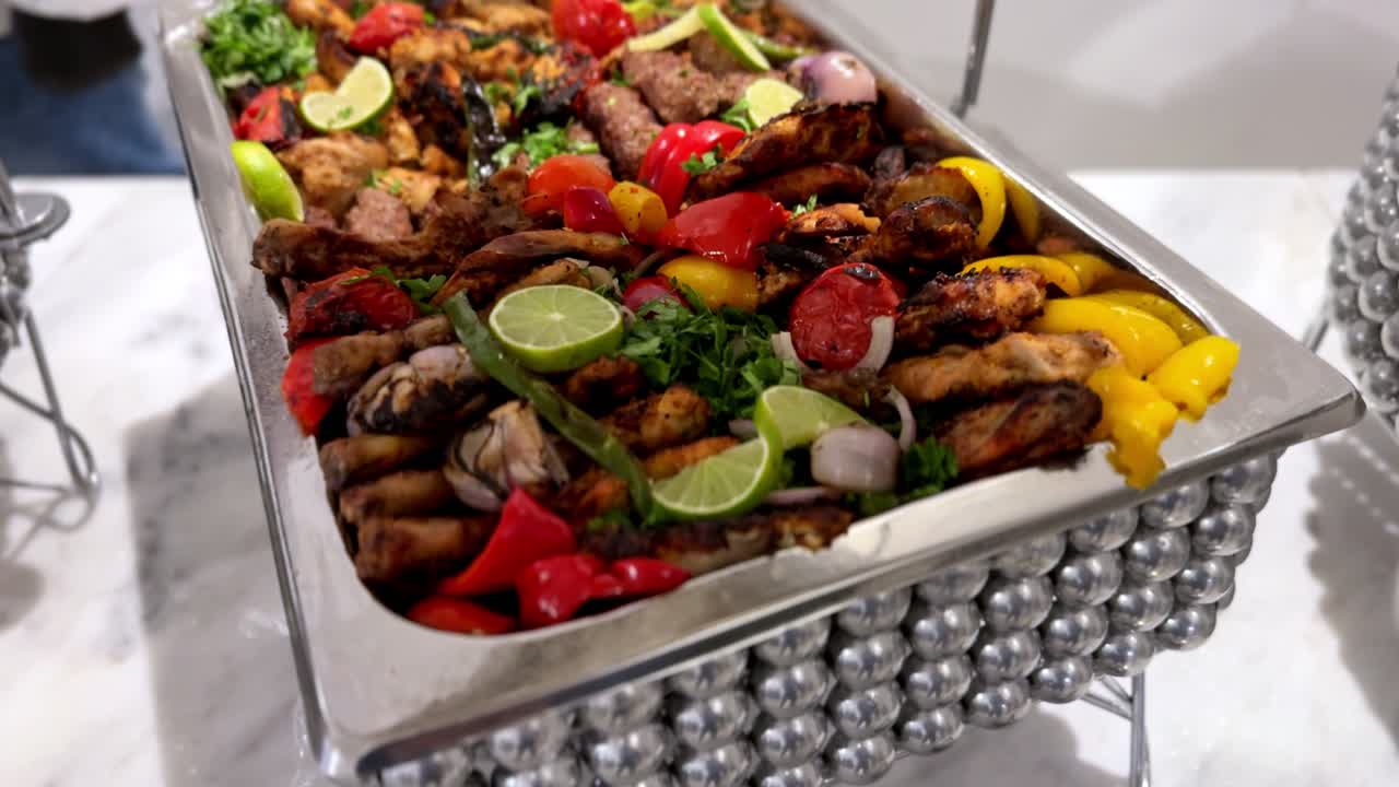 Primo piano di uno scaldavivande in acciaio inossidabile ripieno di spiedini misti alla griglia e peperoni arrostiti, guarnito con erbe fresche e fette di lime, servito a un buffet per banchetti interni sotto illuminazione brillante.