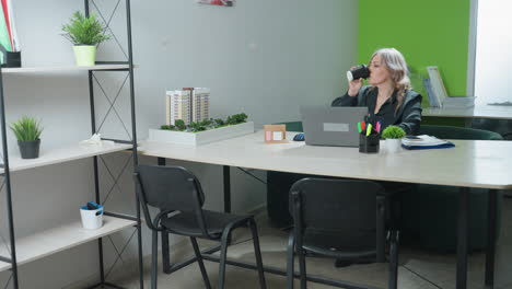 ceo seated at modern office desk pauses focused laptop work to sip latte before resuming duties, surrounded by desk supplies, documents, and architectural model in bright professional workspace