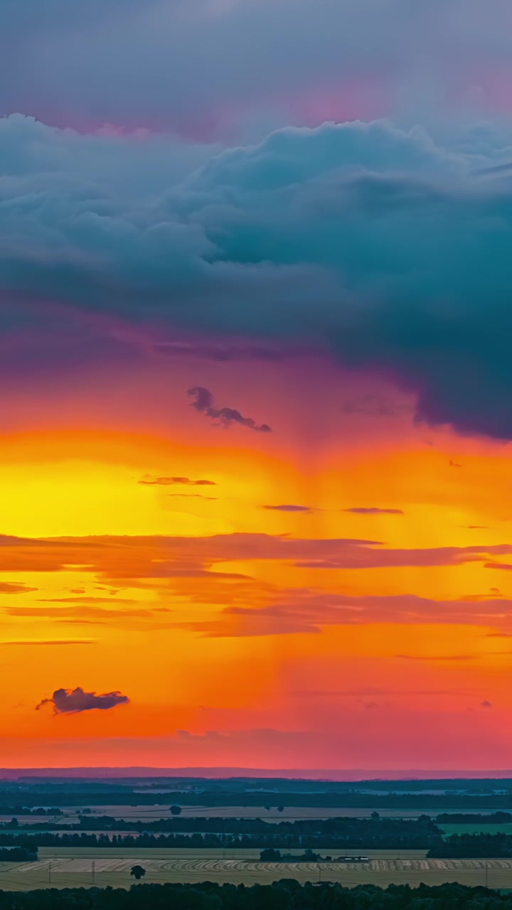 Vivid orange sunset glows beneath heavy storm clouds tinged pink and purple over flat farmland horizon in vertical timelapse