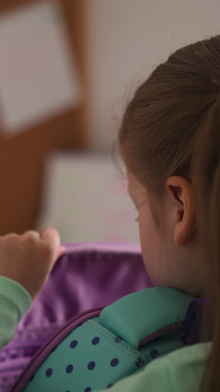Little schoolgirl with ponytails puts textbook from schoolbag on desk at home. Girl prepares to do homework tasks at table after school classes