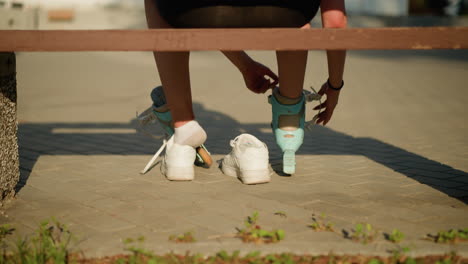 back view of woman strapping roller skate on right leg while other roller skate rests on left leg, with left leg slightly out of sneaker, seated on bench outdoors, with warm sunlight in background