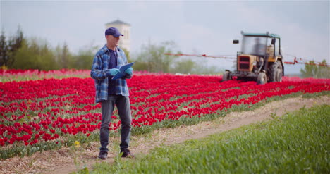 Agricultor-Trabajando-En-El-Campo-Agrícola-Escribiendo-En-Portapapeles-Tractor-Rociando-Tulipanes-Con-Herbicidas