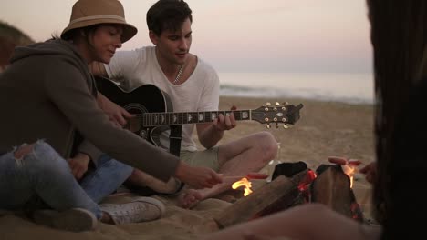 young man is playing guitar by the fire sitting on the beach together with friends. his girlfriend grilling a sausage for him