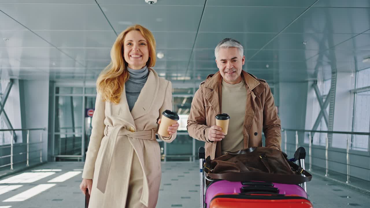 Coppia di viaggiatori maturi e felici che si godono un caffè da asporto, mentre percorrono il terminal dell'aeroporto con un carrello portabagagli