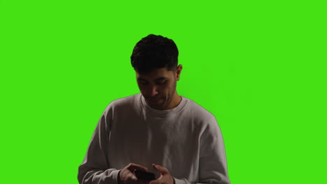 Young-Man-Looking-At-Text-Message-Or-Browsing-Online-On-Mobile-Phone-Standing-Against-Green-Screen-Background-With-Low-Key-Lighting-1