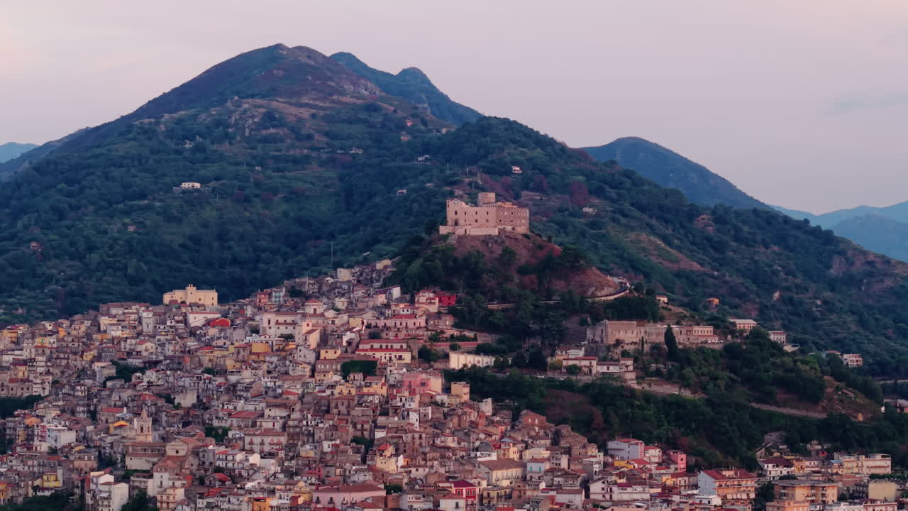 Vista da drone di un borgo collinare siciliano in Italia, circondato da montagne, con edifici storici, case fitte, un'atmosfera tranquilla del centro storico e un paesaggio rurale mediterraneo.