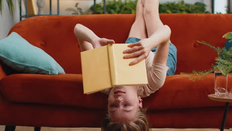 young smiling woman lying upside down on sofa reading interesting book turning pages taking a couch