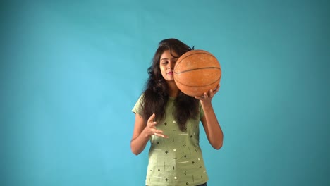A-young-Indian-girl-in-green-t-shirt-playing-with-a-basket-ball-looking-at-the-camera-standing-in-an-isolated-blue-background-studio