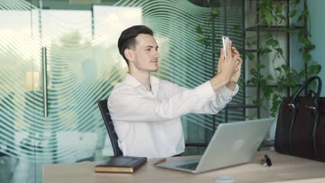 hombre guapo, joven y elegante con ropa de negocios, sentado en un escritorio en una oficina moderna, tomando una selfie con su teléfono inteligente