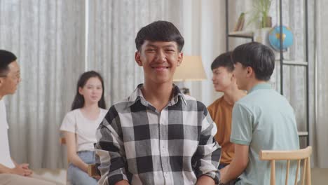 close up of a boy in plaid shirt smiling to camera. asian teen group sitting in chairs forming a circle discussing at home