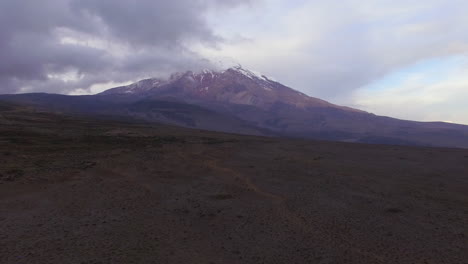 Toma-Aérea-Que-Se-Eleva-Desde-Un-Plano-Volcánico-Para-Mostrar-El-Cotopaxi-Parcialmente-Oculto-Por-Nubes-Oscuras