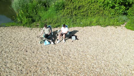 drone footage of two people relaxing on a pebble beach at netley abbey, hampshire. surrounded by greenery and sunshine, they sit in chairs enjoying a peaceful, sunny moment by the water.
