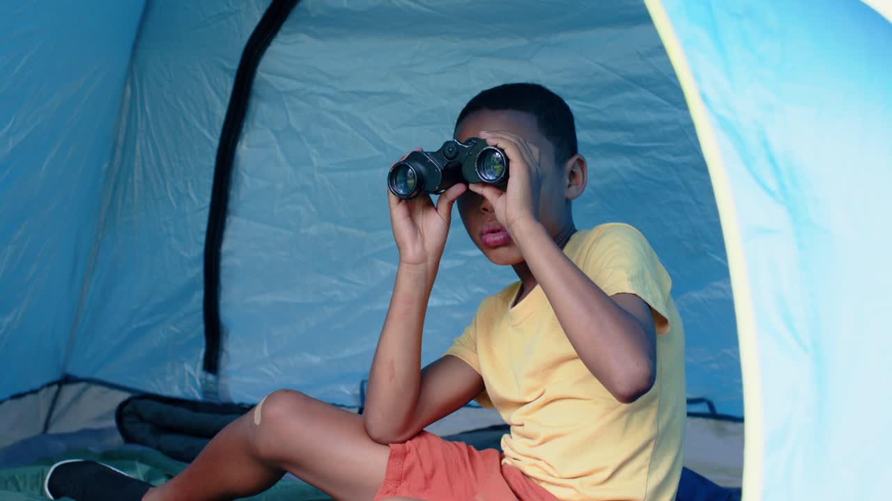 Bambino seduto su un materassino all'interno di una tenda, che alza un binocolo nero mentre avvista un movimento all'esterno