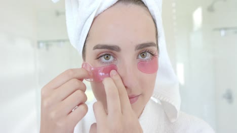 portrait of caucasian woman with towel on head applying eye patches in bathroom in slow motion