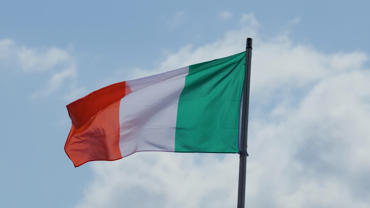 Irish national flag flutters dynamically on flagpole against blue sky and clouds, daylight, outdoor setting