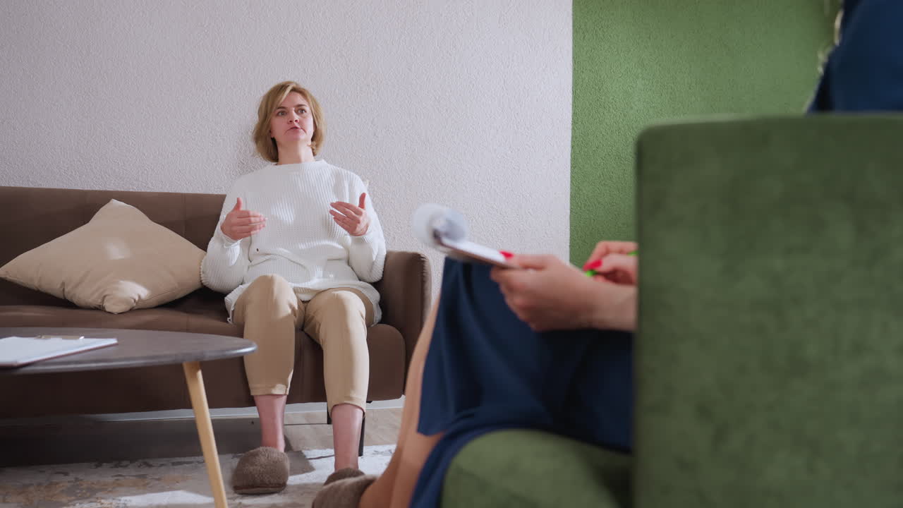Open hearted person demonstrating with hands while speaking to listening partner taking notes in cozy counseling room with brown sofa green accent wall supportive atmosphere clock pen pack visible