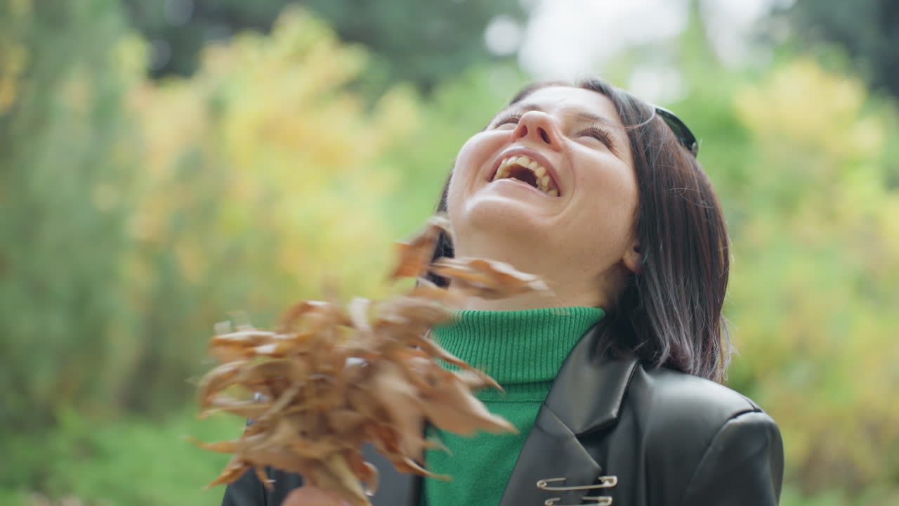 Yapraklı deri ceketli, yeşil kazaklı ve güneş gözlüklü bir kadın, sıcak yapraklar ve yumuşak bokeh ile sonbahar parkında neşeli ve içten bir şekilde gülüyor, kendinden emin ve rahat bir poz veriyor