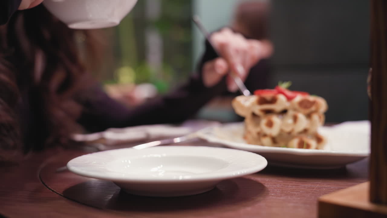 Lady seated at cafeteria table enjoys moment with one hand holding cup near mouth and other grasping fork, close view reveals creamy waffle stack and white ceramic cup on wooden surface in cozy booth