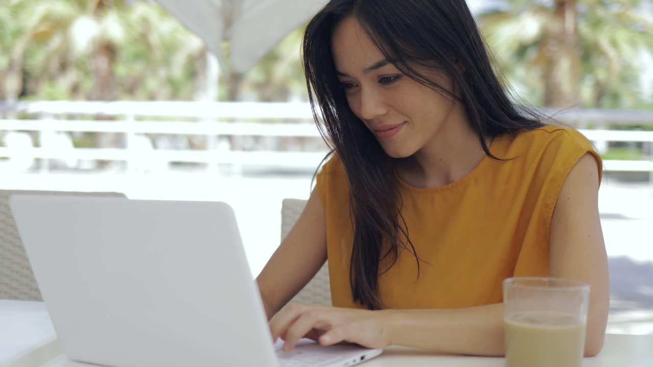 adorable mujer de raza mixta sentada en la mesa de la cafetería al aire libre y usando una computadora portátil