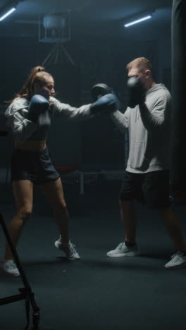 vertical shot of female boxer in boxing gloves hits punching mitts and practices fighting techniques with coach in dark boxing gym. athletic woman exercises and prepares for match.