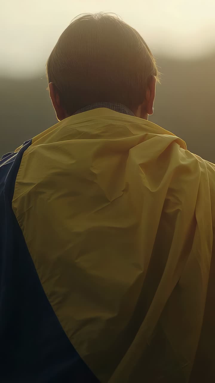 Vídeo vertical: hombre de pie en una colina mirando a la puesta de sol, capa amarilla con panel azul ondeando en la brisa