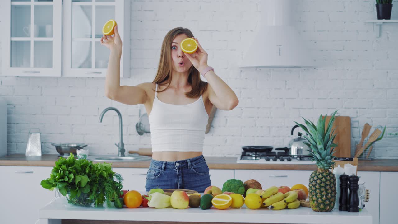 Young woman with oranges in the kitchen and many fresh fruit and vegetables on the table. Beautiful female holding two halves of orange closing her eyes and looking at camera merrily.