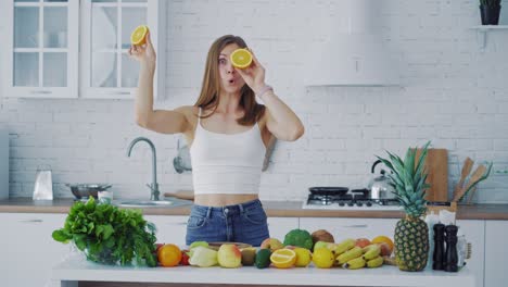 young woman with oranges in the kitchen and many fresh fruit and vegetables on the table. beautiful female holding two halves of orange closing her eyes and looking at camera merrily.
