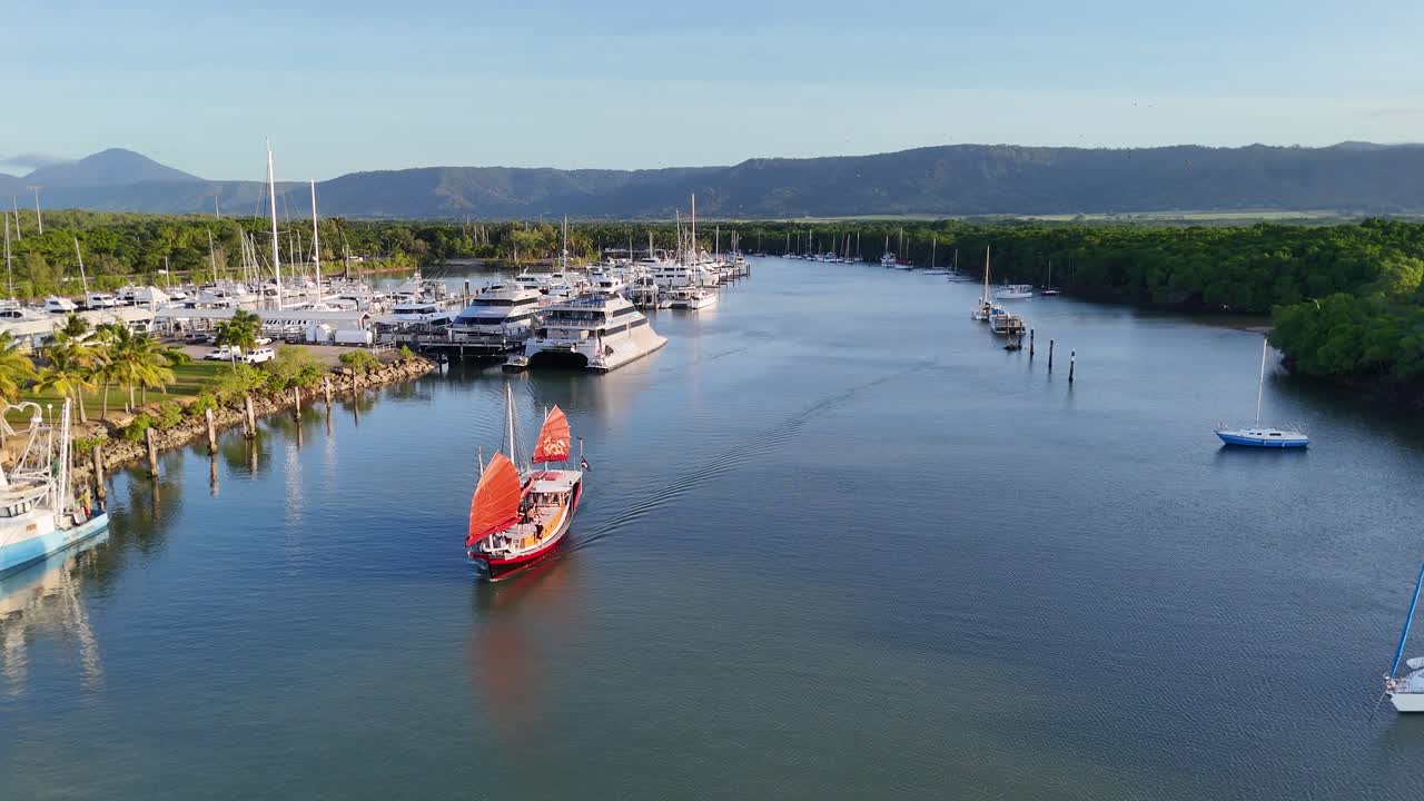Aerial footage captures a red-sailed tour boat cruising into a marina surrounded by yachts and lush greenery, under soft golden hour lighting